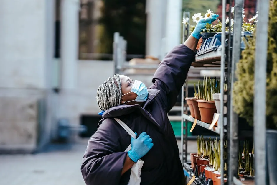 Side view of adult black woman in mask and gloves wearing warm clothes standing in market and choosing potted plants in daytime