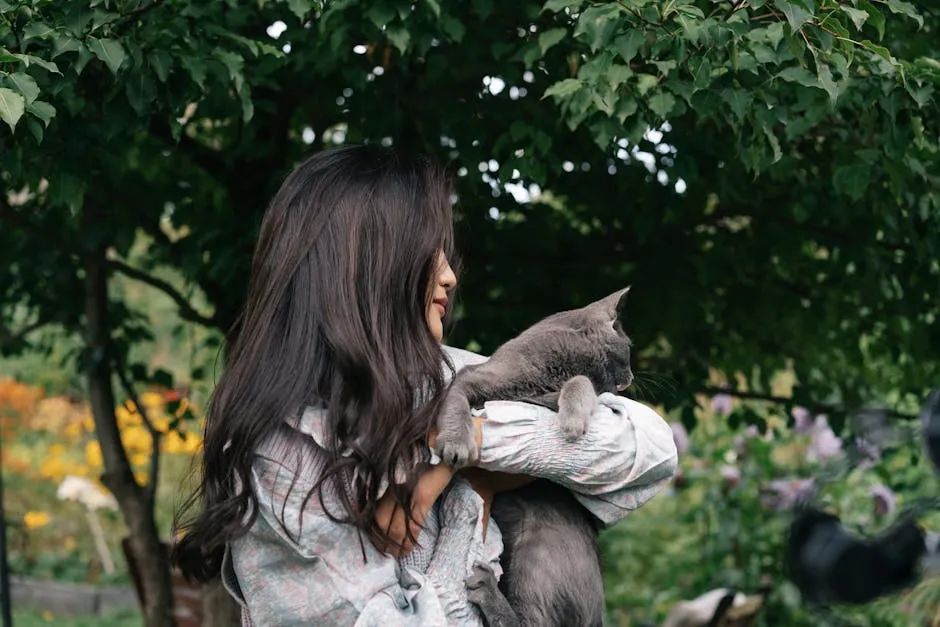 A woman with long hair hugs a gray cat outdoors in a lush garden.