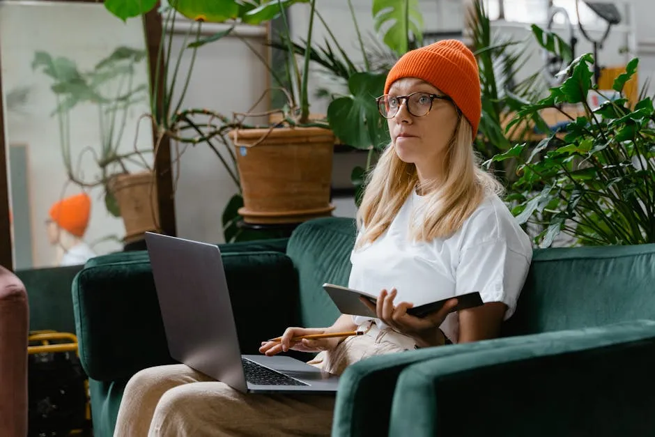 A woman with a notebook and laptop surrounded by plants in a cozy indoor setting.