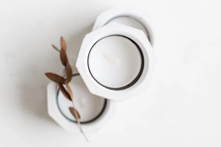 Top view of white candles in minimalist holders placed on marble table with thin stem of dry leaves in sunlight